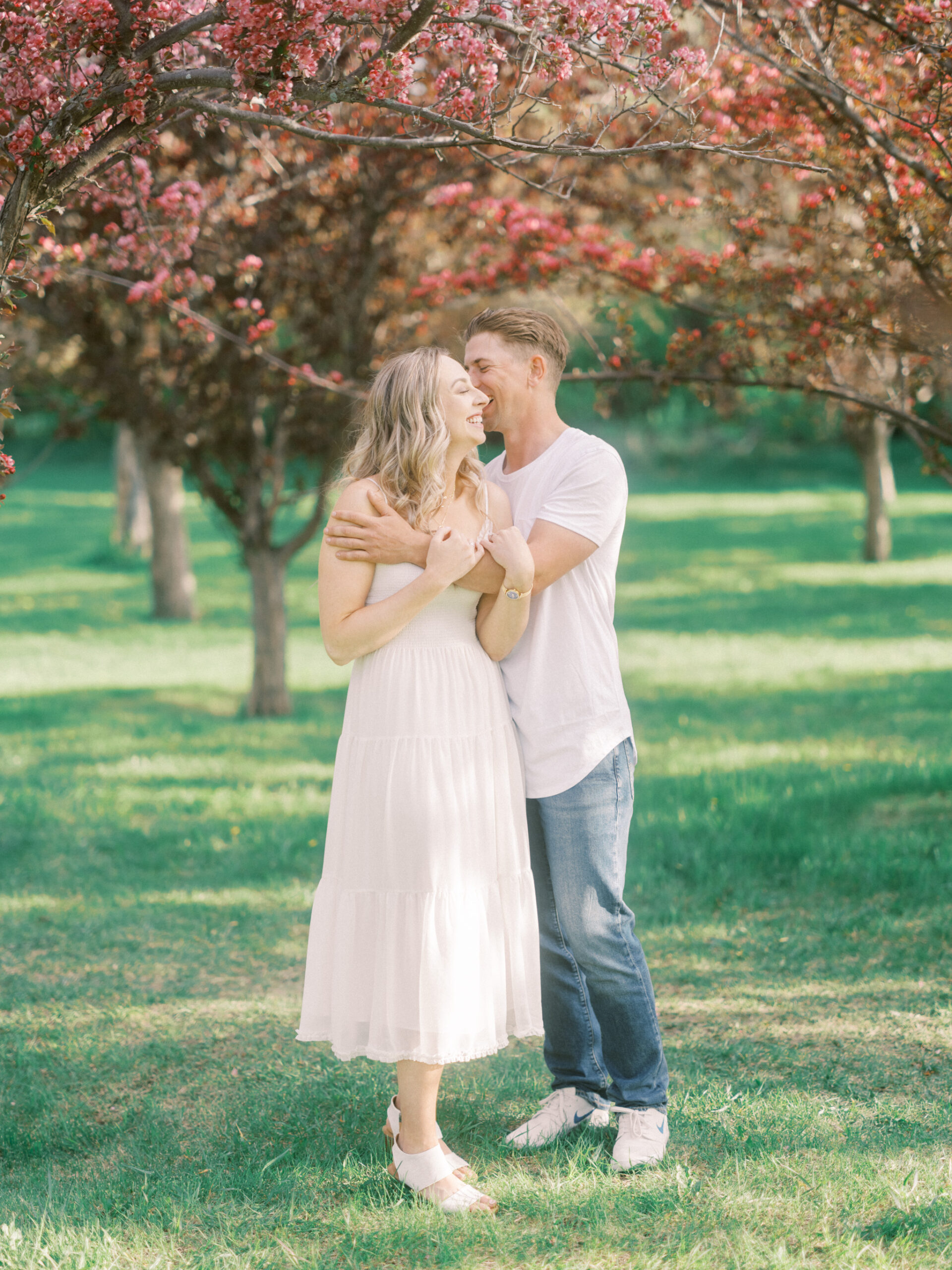 Cherry Blossom Spring Engagement Session, river engagement couple laughing, couple embracing, engagement pose sunset, sunset engagement photos, labrador retriever engagement photos, dog engagement session, puppy engagement session, alberta wedding photographers, alberta photographer, calgary wedding, calgary wedding photographer, spring engagement session, spring photos, nicole sarah, cherry blossom photos, cherry blossom engagement, wedding poses, white dress engagement, sundress white, engagement photos, bow river, puppy engagement photos, neutral engagement photos