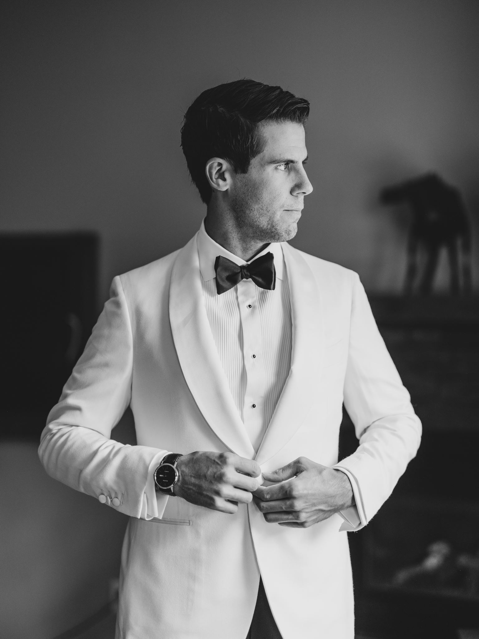 confident groom black and white photo, groom adjusting tuxedo looking out the window, canmore groom white tux, white suit groom with bowtie, The Ultimate Calgary Wedding Photography Guide: Everything You Need to Know Before Your Wedding
