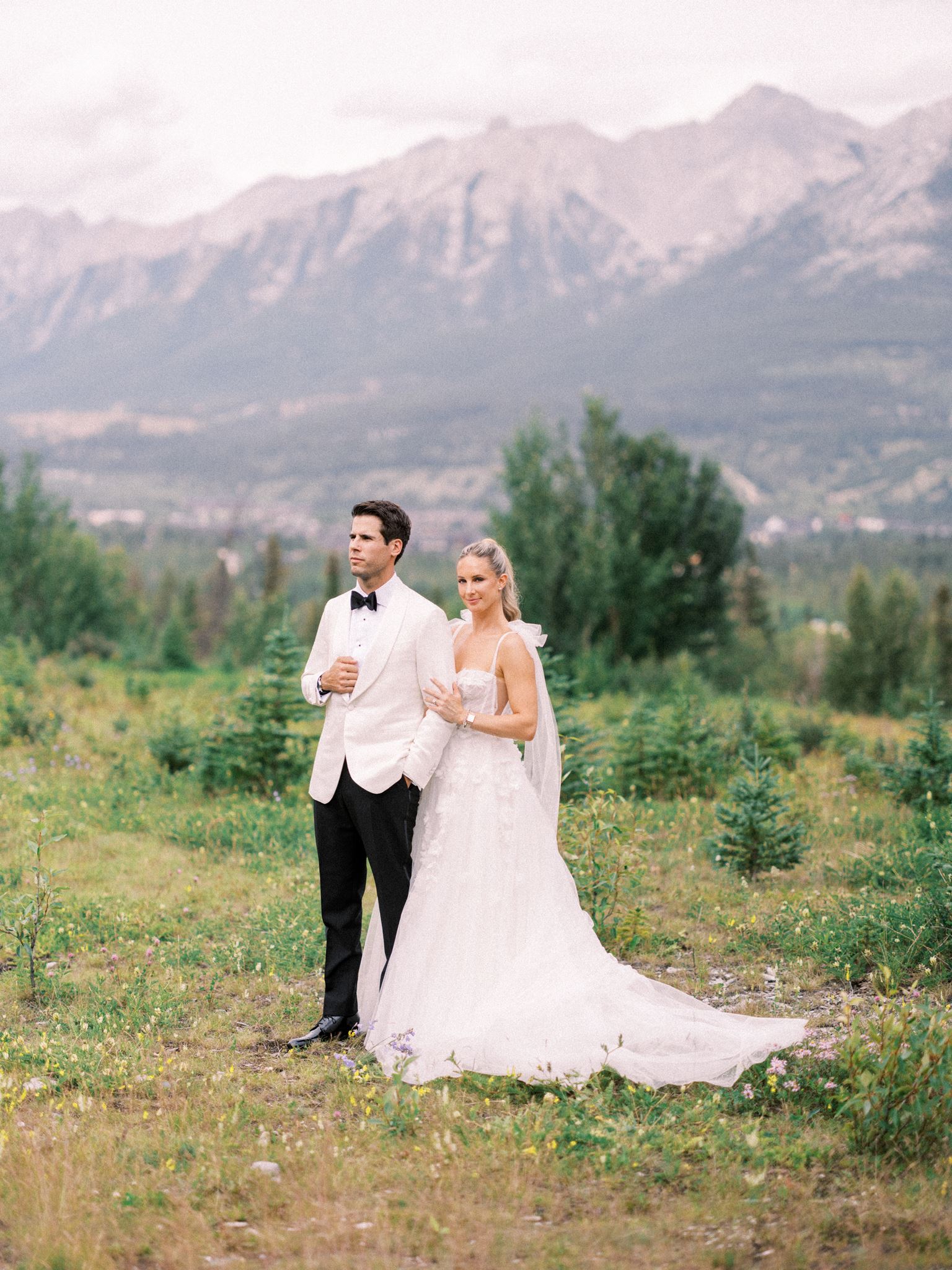 couple in the mountains, bride and groom in canmore, groom in white tuxedo suit, bride in modern wedding dress, groom and bride in a beautiful mountain meadow